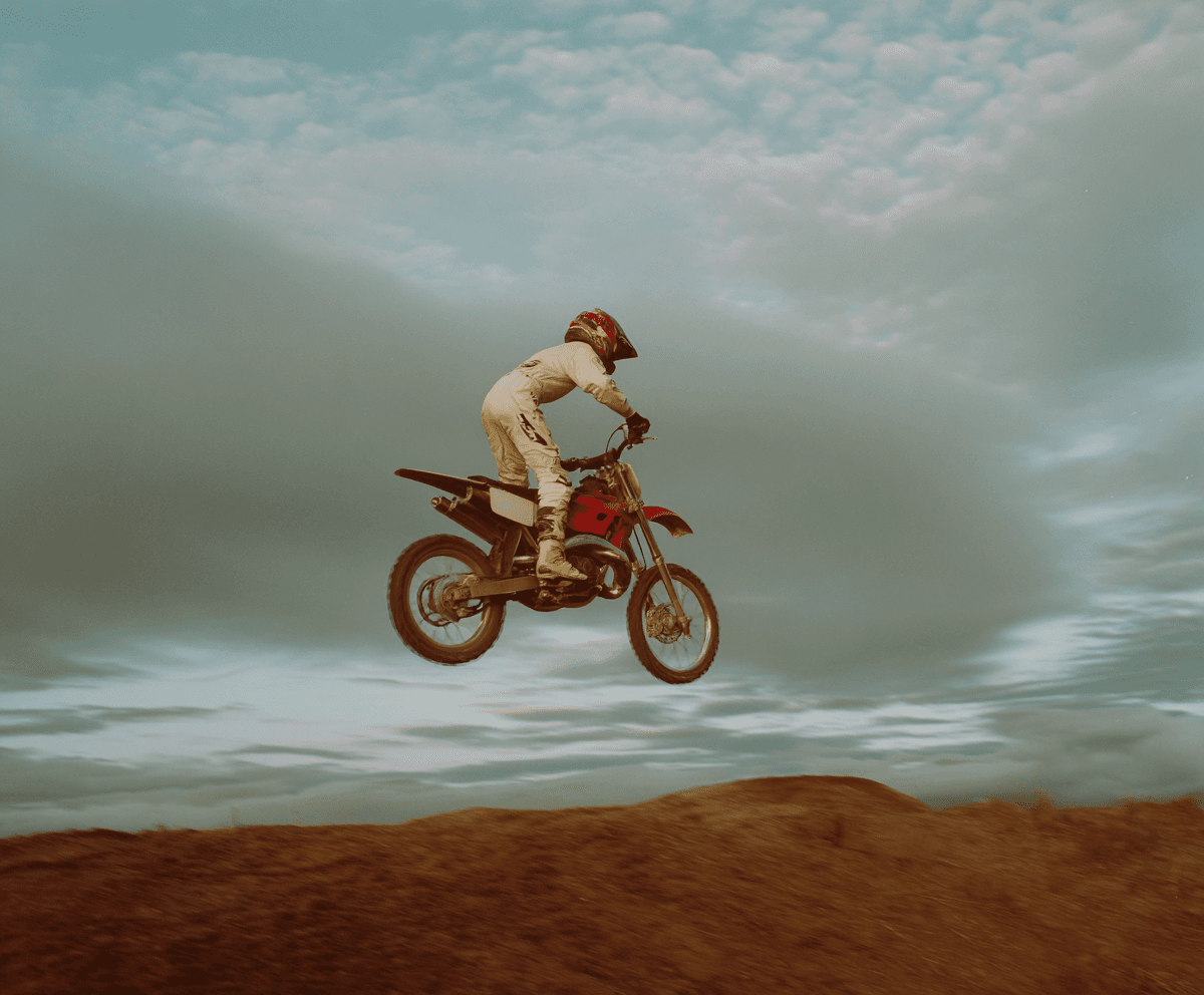 Motocross rider mid-jump above a red-dirt berm under a heavy grey sky